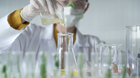 Female scientist researcher wearing a lab coat, white gloves, and mask, is pouring a yellow oily...