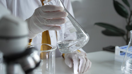 Unknown scientist wearing white protective gloves is carefully shaking up a clear chemical solution inside an Erlenmeyer flask in a laboratory, close up. Science and medicineの写真素材