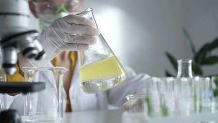 A scientist wearing white protective gloves is shaking up a yellow chemical solution inside an Erlenmeyer flask in a laboratory set, close up. Science and medicine conceptsの写真素材
