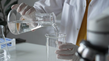 Unknown scientist with a lab coat and white protective gloves is carefully pouring a liquid from a...