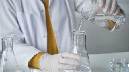 Unknown scientist with a lab coat and white protective gloves is carefully pouring a liquid from a...