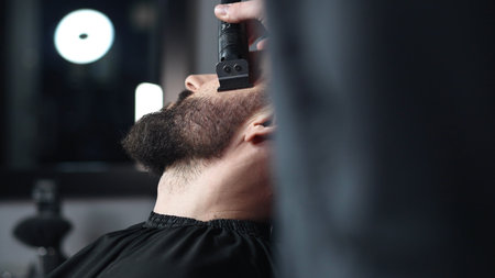 Professional barber trimming clients beard with electric razor, delivering precise styling within contemporary grooming salon settingの写真素材