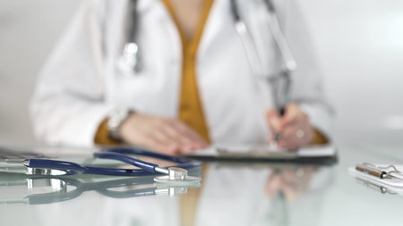 Stethoscope and medication history records form are on a glass table before a female doctor in yellow blouse under a white medical coat. Physician is working on clinical office background. Medicine conceptの写真素材