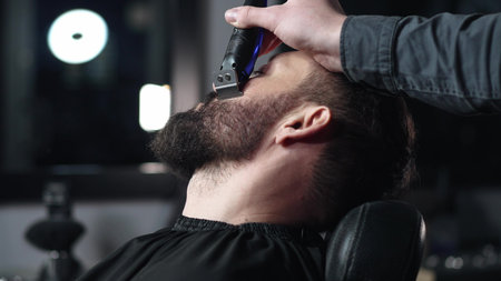 Professional barber trimming clients beard with electric razor, delivering precise styling within contemporary grooming salon settingの写真素材