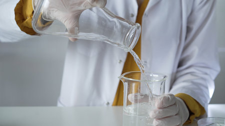 Unknown scientist wearing a lab coat and gloves is carefully pouring a liquid from one beaker into another, close up. Science and medicine conceptsの写真素材