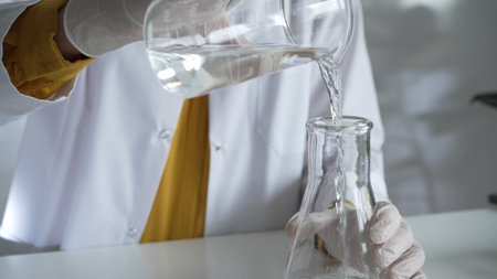 Unknown scientist wearing a lab coat and gloves is carefully pouring a liquid from a beaker into an...