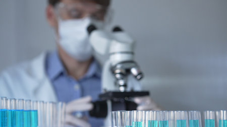 Laboratory tubes are in focus, closeup view. Man scientist researcher with a white protective gloves is using a microscope at the background. Medicine and scienceの写真素材
