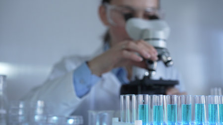 Laboratory tubes, filled with a blue liquid, are in focus, closeup view. Female scientist researcher is using a microscope at the backgroundの写真素材