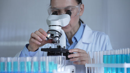 A female scientist, wearing a lab coat and safety glasses, is adjusting a binocular microscope surrounded by test tubes in laboratory setting. Medicine and scienceの写真素材