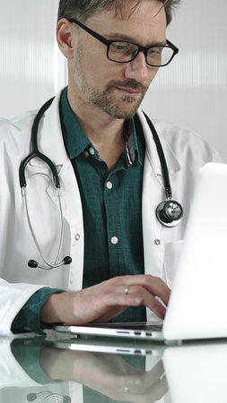 A male doctor in a green shirt under a medical coat is using technology in healthcare. Man is typing on a laptop on a glass desk in the clinic, vertical portrait view. Medicineの写真素材