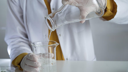Unknown scientist wearing a lab coat and gloves is carefully pouring a liquid from one beaker into another, close up. Science and medicine conceptsの写真素材