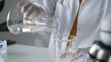 Unknown scientist wearing a lab coat and gloves is carefully pouring a liquid from a beaker into an Erlenmeyer flask, close up. Science and medicineの写真素材