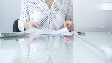 Businesswoman in white dotted blouse is going through paperwork while holding documents at a glass desk in a well lit, contemporary office setting. Business people conceptの写真素材