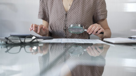 Businesswoman accountant in dotted blouse reviewing financial reports with magnifying glass and calculator, conducting audit and analyzing data in office. Taxes, audit in businessの写真素材
