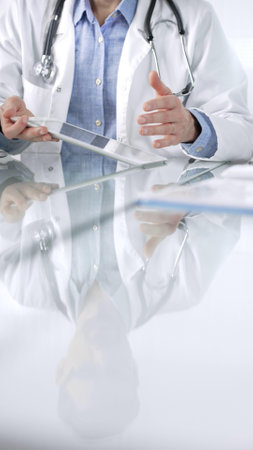 Unknown male doctor wearing a stethoscope, blue shirt and a lab coat is gesturing with hands overs a glass desk with digital tablet in clinic office, vertical closeup view. Medicine conceptの写真素材