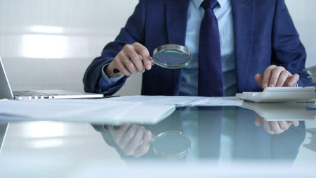 Businessman analyzing financial data with magnifying glass and calculator, conducting audit or review in office. Audit and taxes theme in businessの写真素材