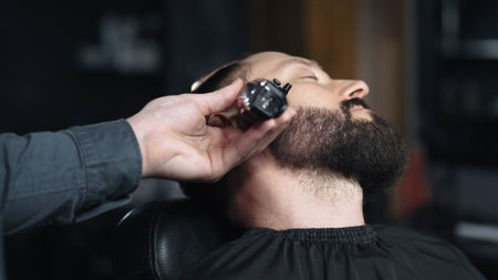 Professional barber trimming clients beard with electric razor, delivering precise styling within contemporary grooming salon settingの写真素材