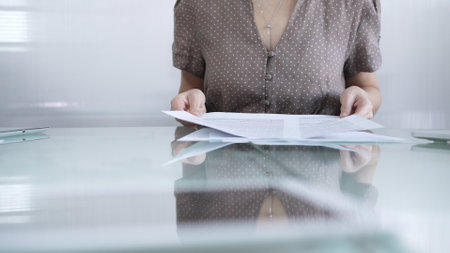 Businesswoman or accountant in dotted blouse is going through paperwork while holding documents at a glass desk in a well lit, contemporary office setting. Business people conceptの写真素材