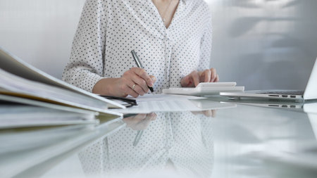 Focused businesswoman at a sleek glass desk in a contemporary office, using a calculator to crunch...