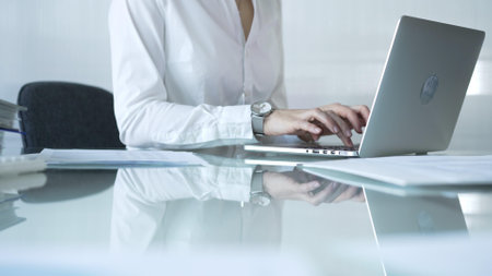 Businesswoman in white blouse typing on laptop at glass desk while sitting in bright office, showcasing productivity and modern work environment. Businesspeople conceptの写真素材