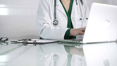 Female doctor using technology in healthcare, close up of hands typing on laptop with stethoscope, glasses, and clipboard on deskの写真素材