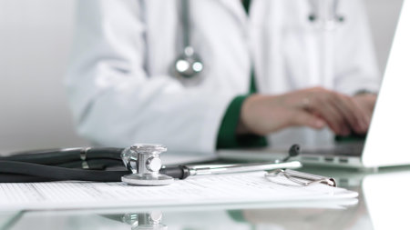 Stethoscope and medication history records form are lying on a glass table before a doctor working with laptop in clinical office background, close-up view. Medicine conceptの写真素材
