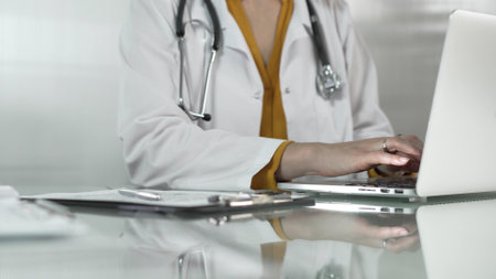 Female doctor, wearing yellow blouse under a medical coat, is using technology in healthcare, close up of hands typing on laptop with stethoscope, glasses, and clipboard on desk. Medicine conceptの写真素材