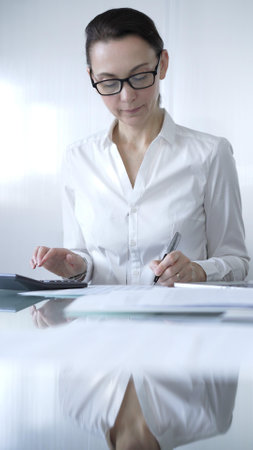 Businesswoman or accountant, wearing white casual blouse and glasses, is working on tax calculations, note taking, and financial reports in the office. Audit and taxes in businessの写真素材