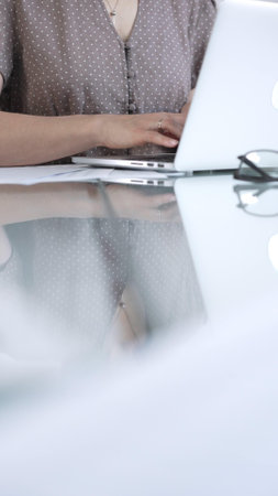 Businesswoman is typing on laptop keyboard, preparing taxes and audit reports, with documents on glass office desk, vertical view. Taxes, audit in businessの写真素材