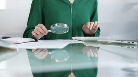 Businesswoman in green blouse carefully reviewing financial reports with magnifying glass and calculator, conducting audit and analyzing data in office. Taxes, audit in businessの写真素材
