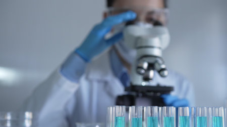 Laboratory tubes, filled with a blue liquid, are in focus, closeup view. Female scientist researcher is using a microscope at the background. Medicine and science conceptの写真素材