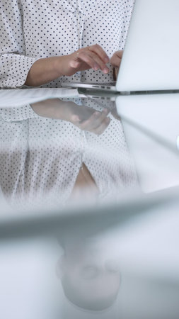 Businesswoman in white dotted blouse is typing on laptop keyboard, preparing taxes and audit reports, with documents and eyeglasses on glass desk, closeup vertical view. Taxes, audit in businessの写真素材
