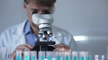A man scientist, wearing a lab coat, mask, and safety glasses, is using a microscope surrounded by test tubes in laboratory setting. Medicine and science conceptの写真素材
