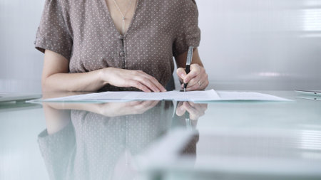 Businesswoman wearing dotted blouse is signing a contract at a sleek glass desk in a modern office, closeup. Business people conceptの写真素材