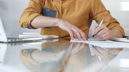 Businessman accountant wearing casual yellow shirt carefully reviewing financial reports using a calculator on office glass desk. Taxes, audit in businessの写真素材