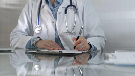 Woman doctor, wearing a blue blouse under a white medical coat, is using a clipboard and taking notes on glass desk in a medical office, close-up. Medicineの写真素材
