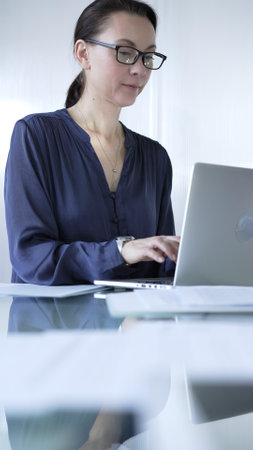 Focused businesswoman with glasses typing on laptop at modern glass desk in well lit office. Business people conceptの写真素材