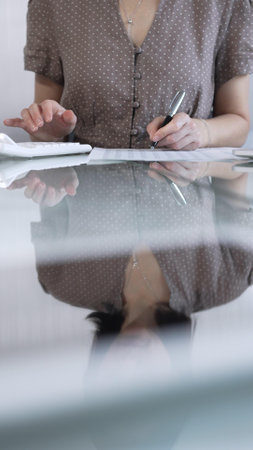 Businesswoman accountant, wearing dotted blouse, is using a calculator at sleek glass desk in a contemporary office, vertical view. Taxes, audit in businessの写真素材
