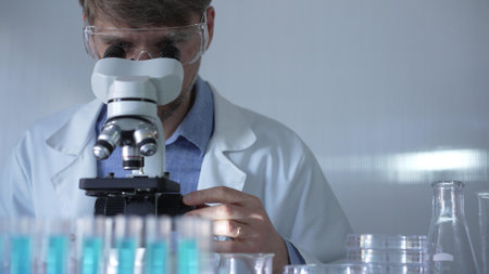 A man scientist, wearing a lab coat, mask, and safety glasses, is using a microscope surrounded by test tubes in laboratory setting. Medicine and science conceptの写真素材