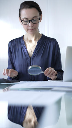Woman accountant examining financial documents with magnifying glass at glass office desk, conducting thorough analysis of business transactions. Audit and taxes in businessの写真素材