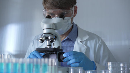 A scientist man, wearing a lab coat, mask, blue gloves and safety glasses, is using a binocular microscope surrounded by test tubes in laboratory. Portrait view. Medicine and scienceの写真素材