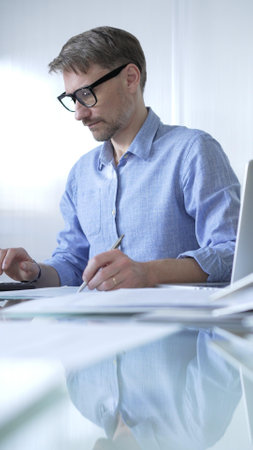 Businessman or accountant, wearing blue casual shirt and glasses, is working on tax calculations, note taking, and financial reports in the office. Audit and taxes in businessの写真素材