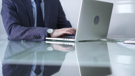 Businessman in blue jacket is focused on typing on laptop at glass desk in modern office, showcasing productivity and concentration, closeup view. Business people conceptの写真素材