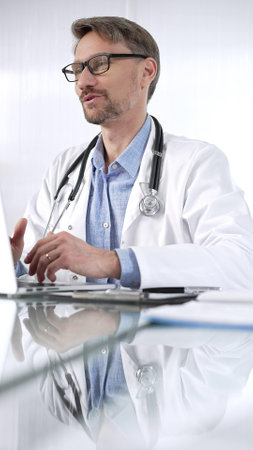 Doctor man, wearing a stethoscope, blue shirt and a lab coat is gesturing with hands overs a glass desk with laptop in clinic office, vertical portrait view. Medicine and scienceの写真素材