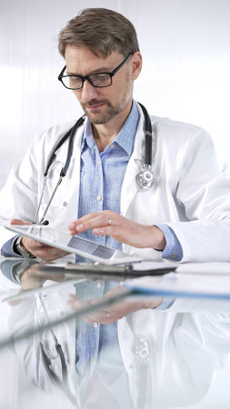 Doctor man in blue shirt under a white coat, glasses and stethoscope is using digital tablet in modern healthcare setting, interacting with patient data, vertical portrait view. Medicine and scienceの写真素材