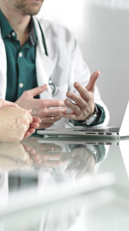Doctor man wearing a stethoscope and lab coat is gesturing with hands while explaining a diagnosis to a patient across a desk with a laptop. Medicine conceptの写真素材