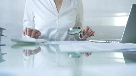 Businesswoman analyzing financial data with magnifying glass and calculator, performing accounting and auditing tasks in office. Audit and taxes theme in businessの写真素材