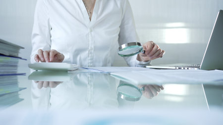 Businesswoman analyzing financial data with magnifying glass and calculator, performing accounting and auditing tasks in office. Audit and taxes theme in businessの写真素材