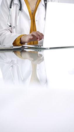 Unknown doctor woman, wearing a stethoscope and yellow blouse under a lab coat, is using technology in healthcare. Close-up of hands typing on a laptop on a glass desk, vertical view. Medical conceptの写真素材