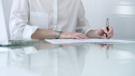 Businessman in white casual shirt is signing a contract at a sleek glass desk in a modern office, showcasing professionalism and focus in his work environment. Business people conceptの写真素材